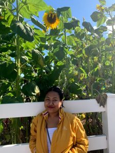 Grace smiling in front of a white fence with tall sunflowers behind her